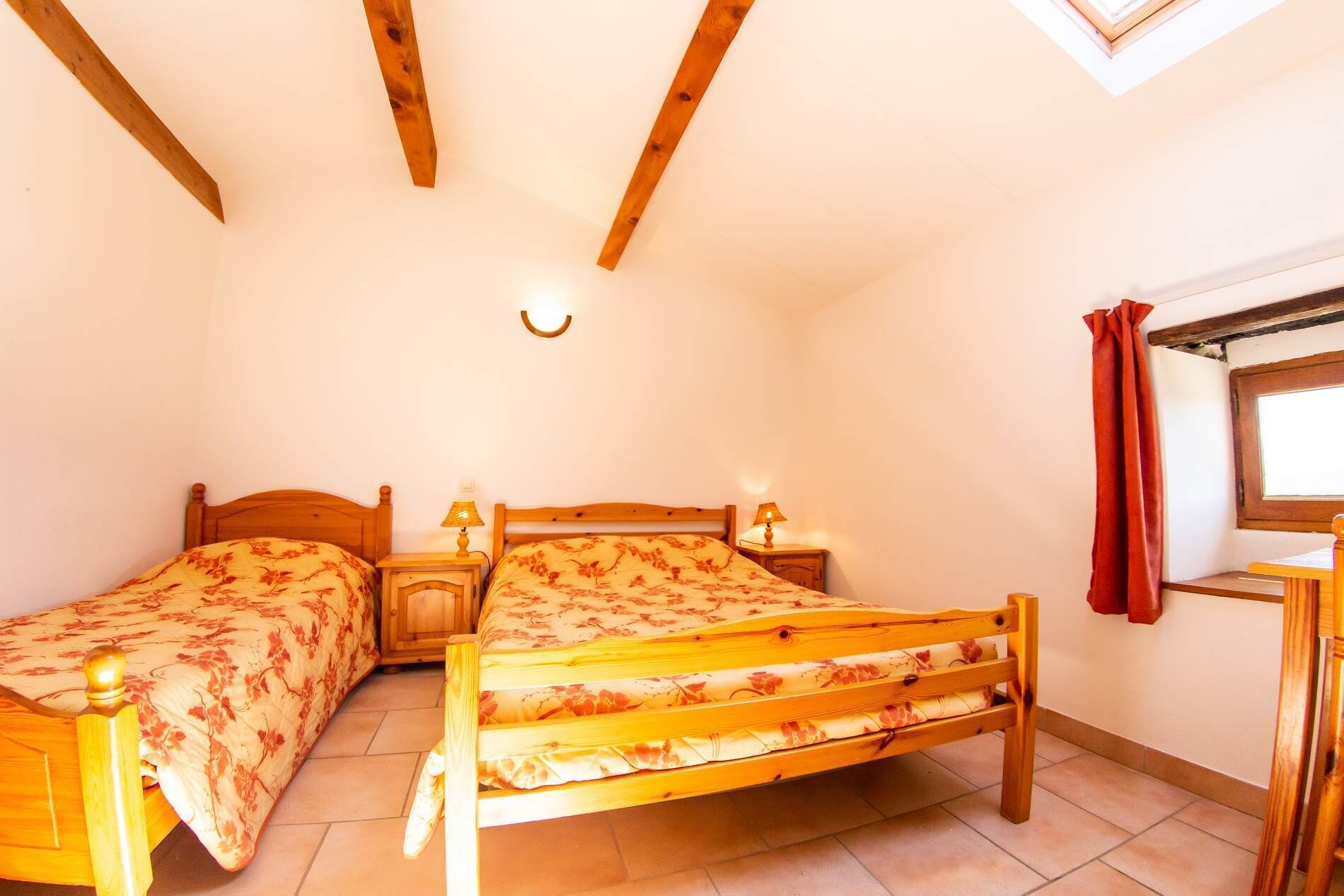 A bedroom with two wooden beds, a small table, a nightstand, a wall-mounted lamp, and a window with a curtain.