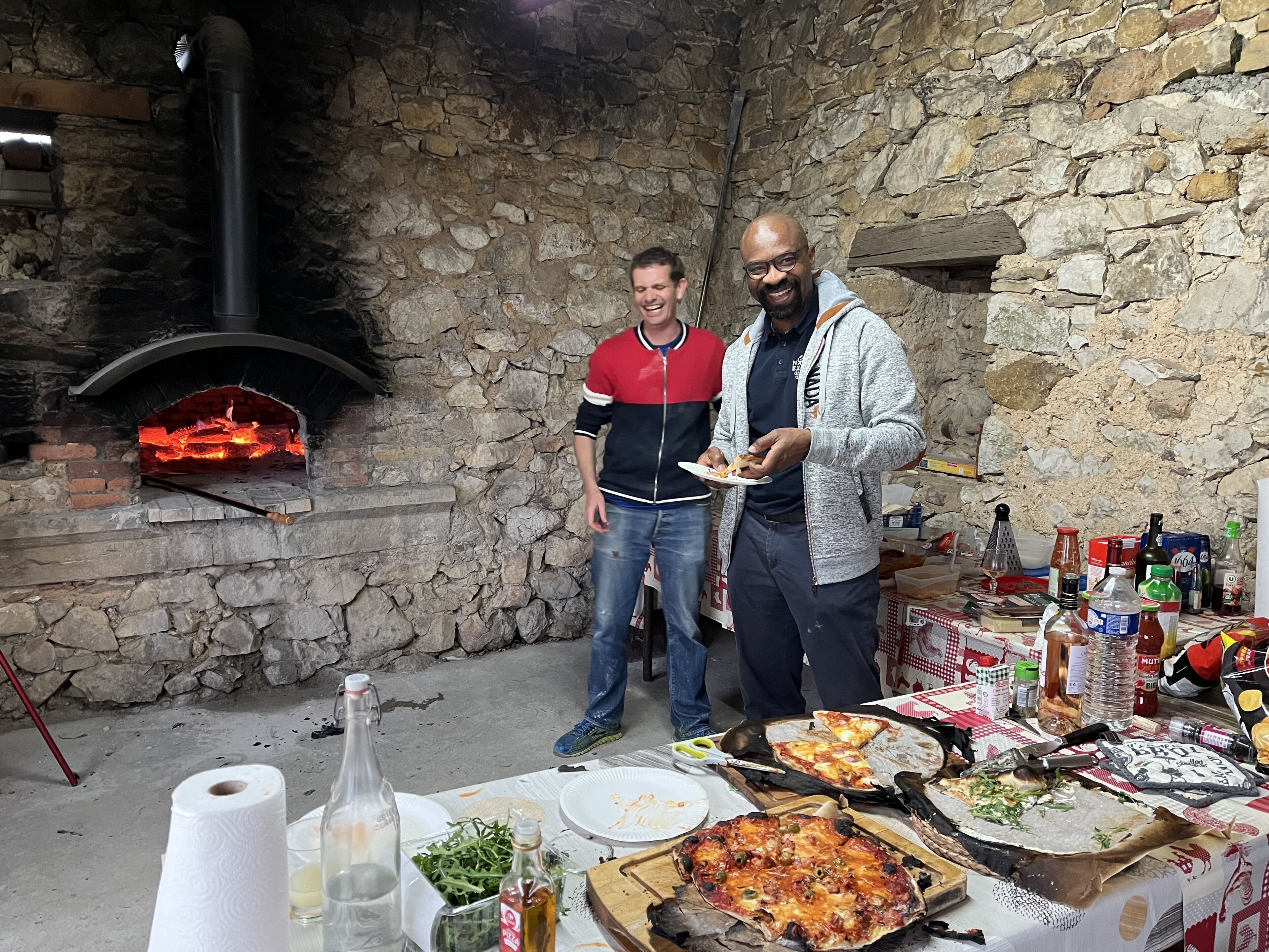 Un espacio al aire libre con paredes de piedra, un horno de ladrillo, pizza y diferentes alimentos sobre una mesa.