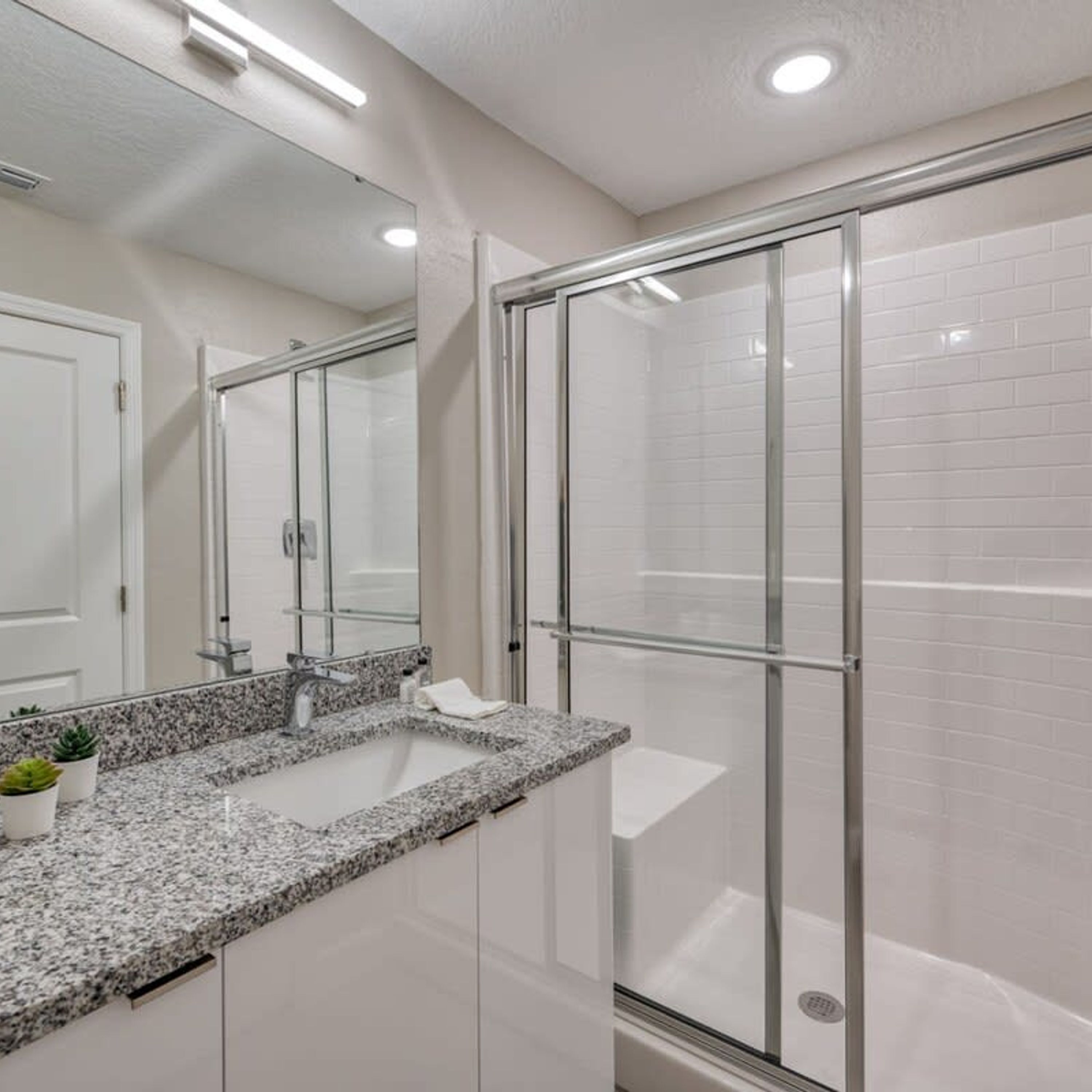 A modern bathroom with a granite countertop, a glass shower enclosure, and white tiled walls.