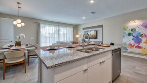 A modern kitchen with a granite countertop, stainless steel sink, and a dishwasher. There is a dining area with a table and chairs, and a large window providing natural light. - Super Spacious Villa With Private Pool and Themed Bedrooms (Kissimmee)