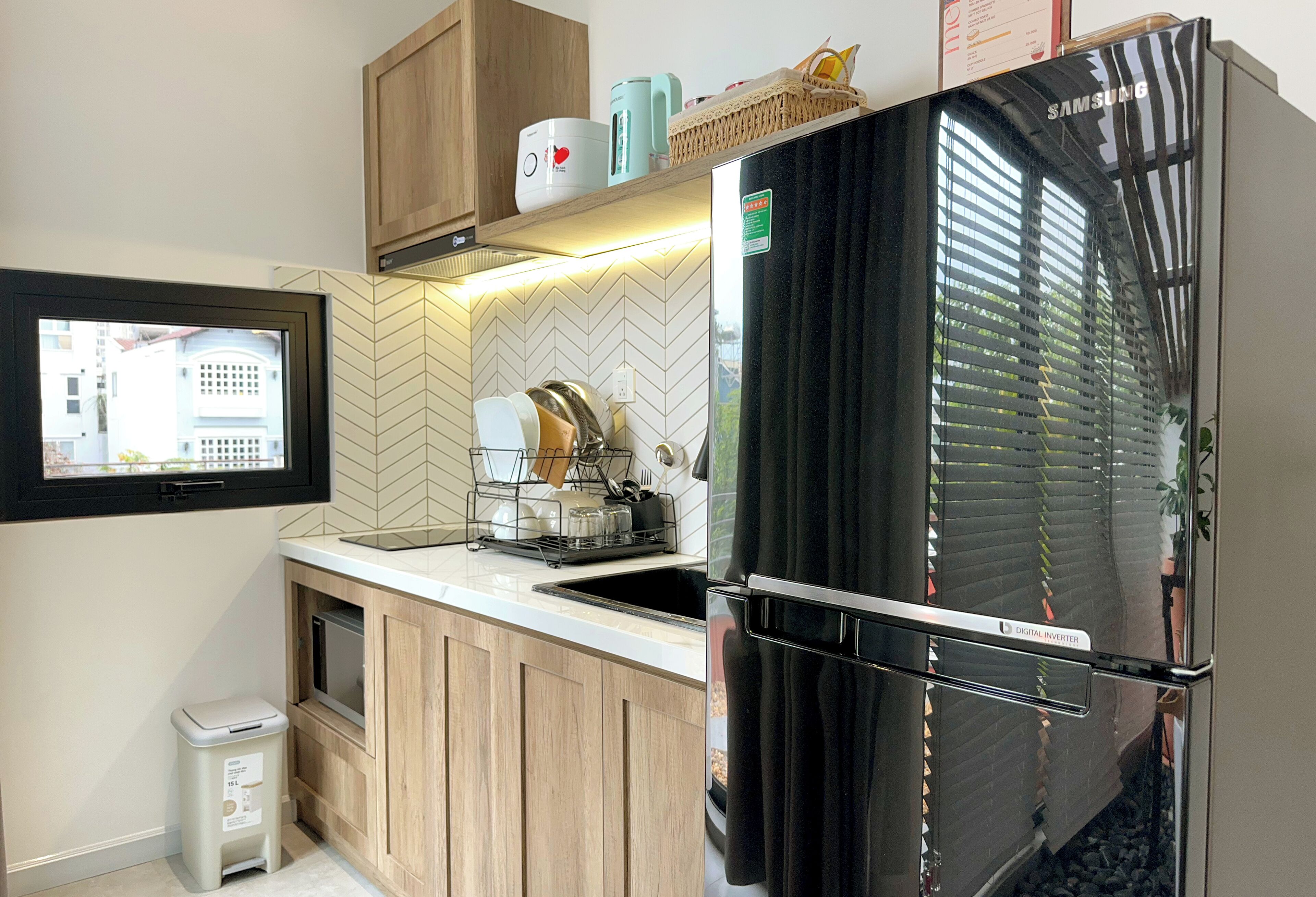Cocina compacta con refrigerador negro, armarios de madera, salpicadero con motivo de espiga y ventana con vista a un edificio.