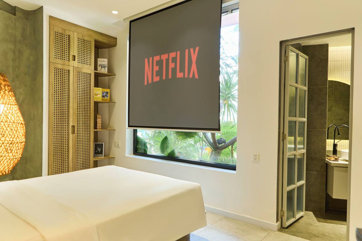 Une chambre avec une grande fenêtre, un lit, une armoire et un écran fixé au mur qui affiche le logo « NETFLIX ».