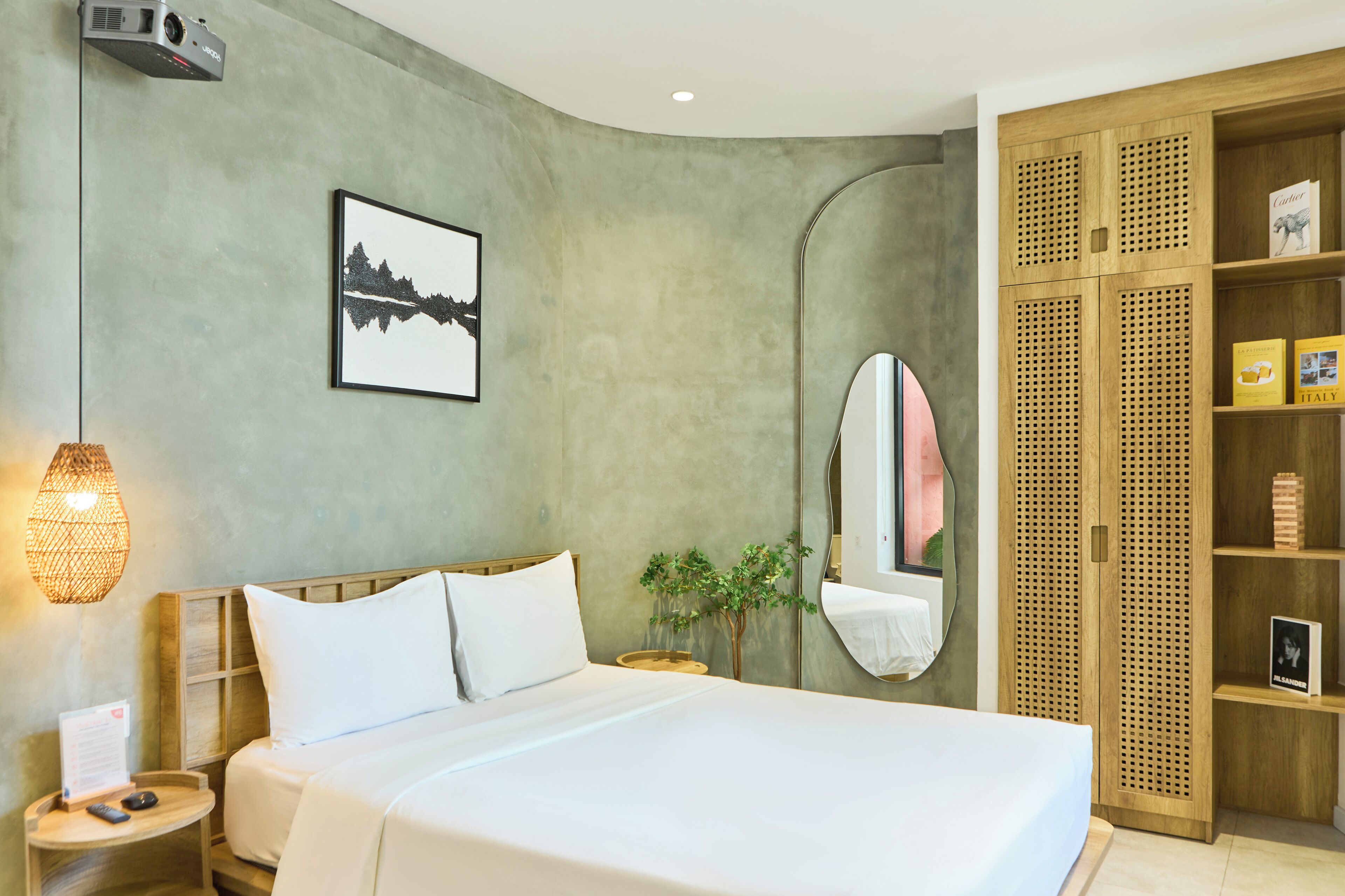 Una camera d'albergo moderna con un letto grande, un comodino con una lampada, una piccola pianta, un quadro appeso e un armadio in legno con ripiani.