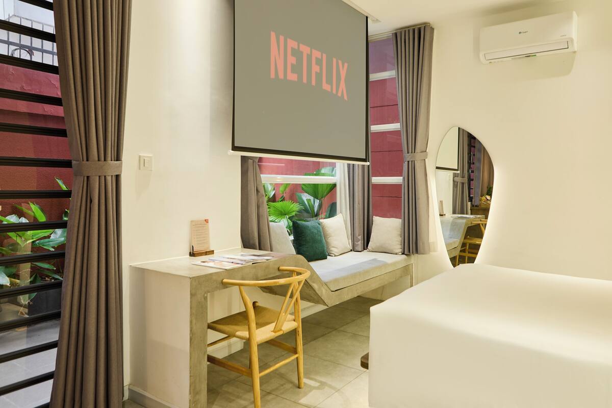 Une chambre moderne équipée d’un bureau, d’une chaise et d’une grande fenêtre avec des rideaux. Un téléviseur à écran plat est fixé au-dessus du bureau et affiche le logo de Netflix.