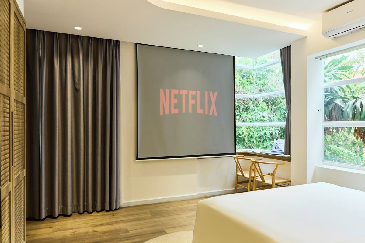 Une chambre d’hôtel moderne dotée d’une grande fenêtre, d’un lit, d’un bureau et d’un écran fixé au mur affichant le logo de Netflix.