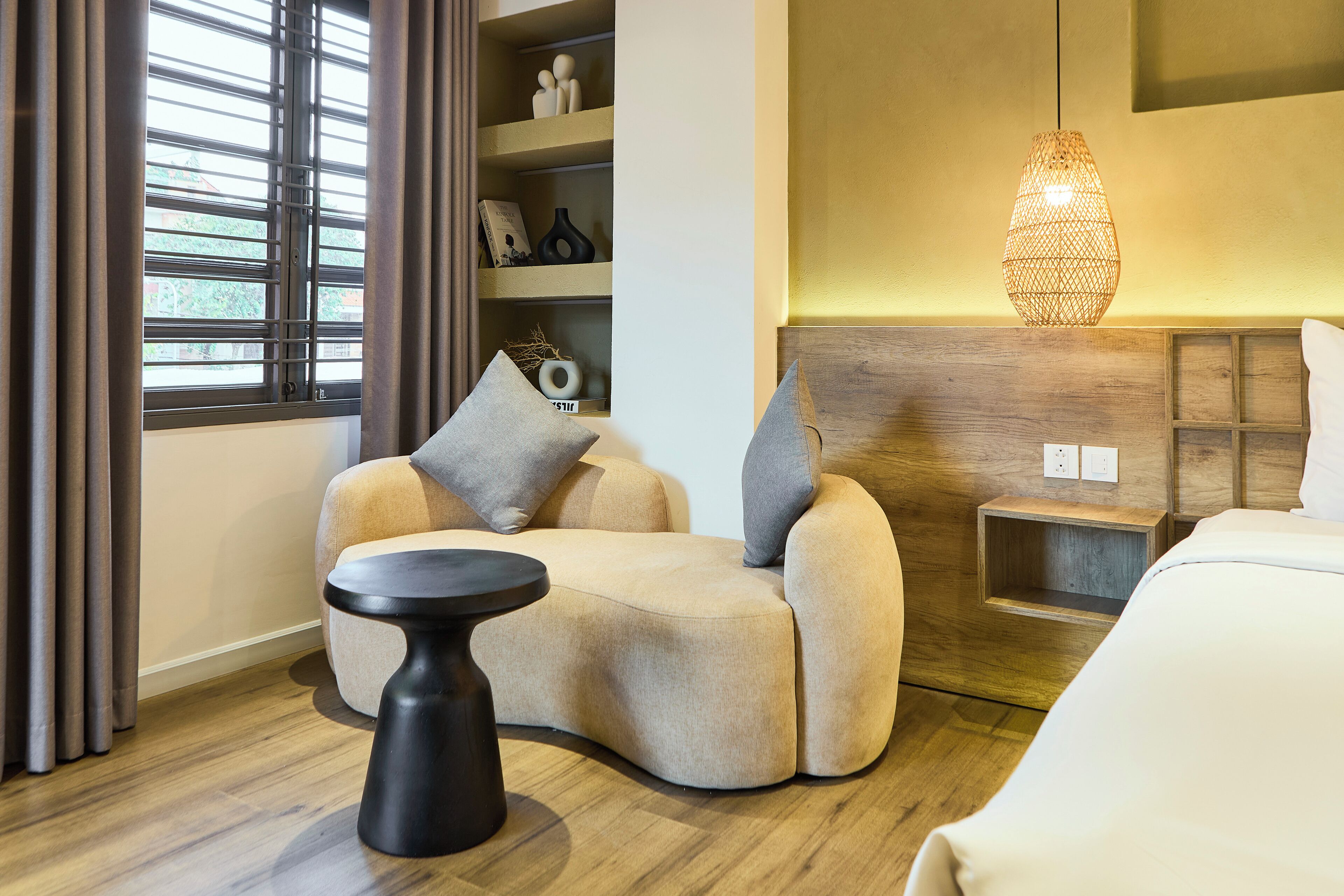 Une chambre d’hôtel moderne avec un canapé incurvé, une petite table ronde, une table de chevet, une tête de lit en bois et un luminaire suspendu.