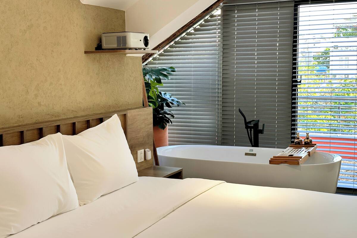 Une chambre d’hôtel moderne avec un grand lit, un projecteur intégré, une grande fenêtre avec stores, une plante en pot et une baignoire avec un plateau de produits de toilette.