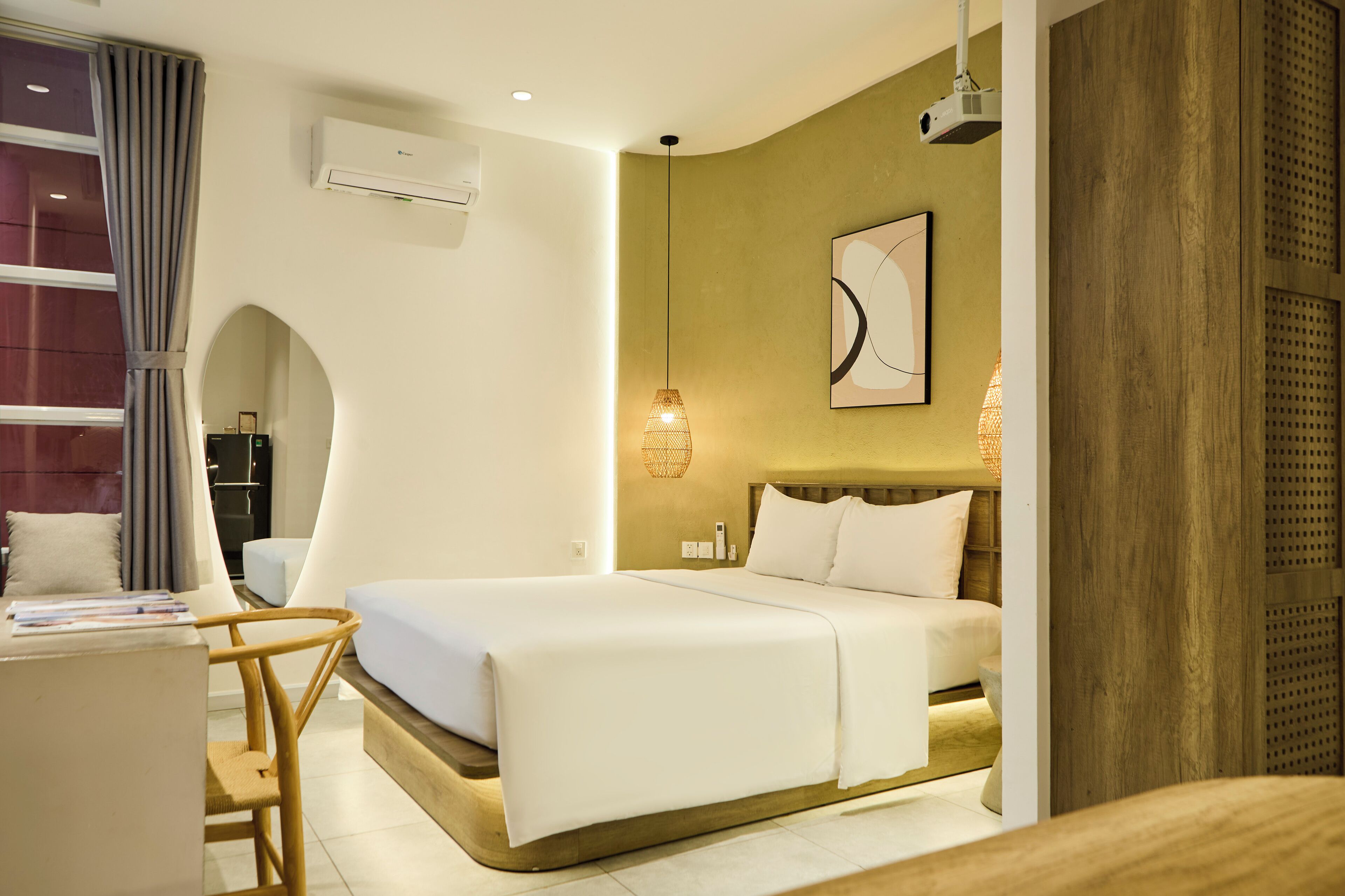 A modern hotel room with a large bed, a desk with a chair, a wall-mounted air conditioner, and a unique arched window.