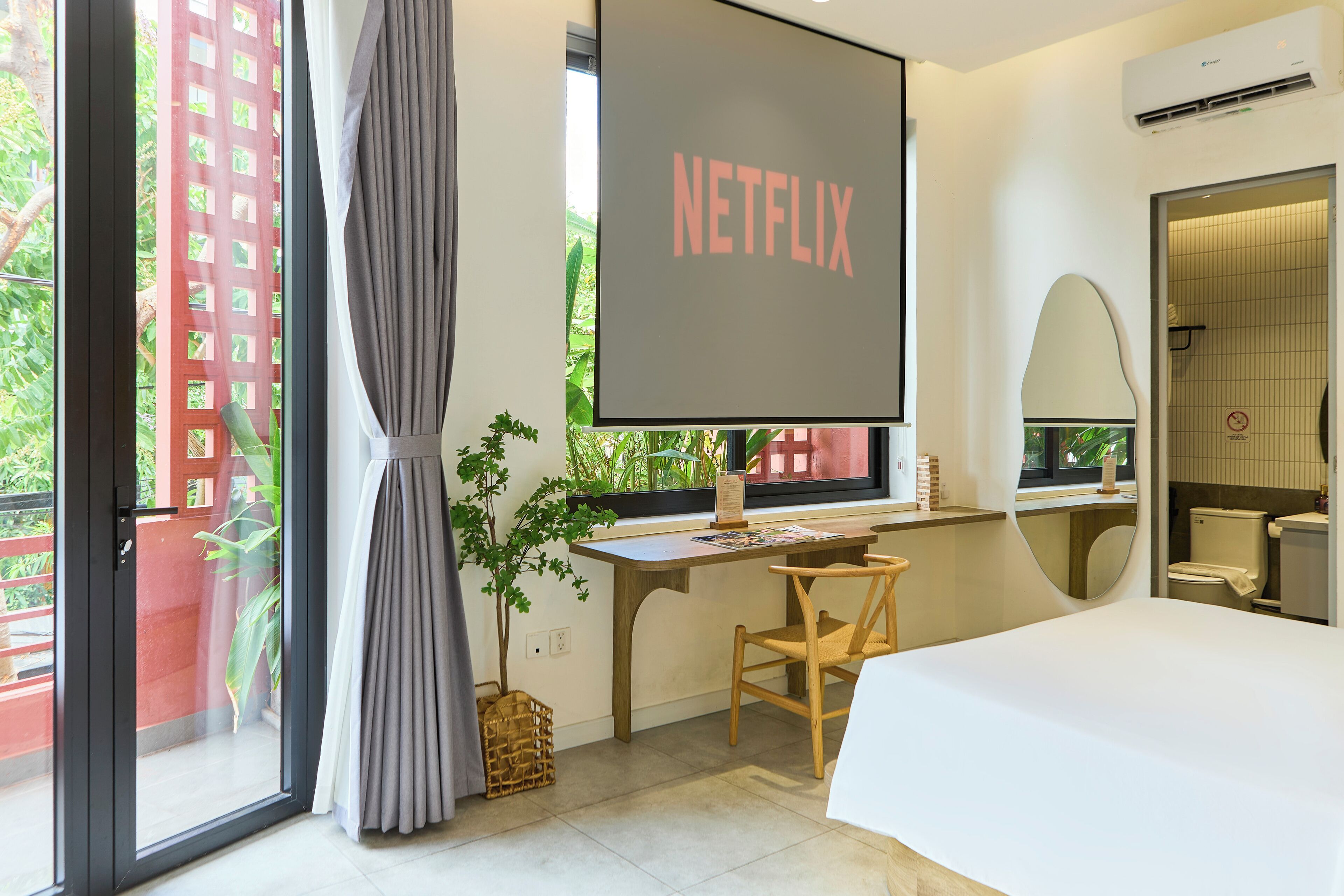 A modern hotel room with a large bed, a desk with a chair, a TV screen displaying "NETFLIX", a window with curtains, a potted plant, and a bathroom visible through the window.