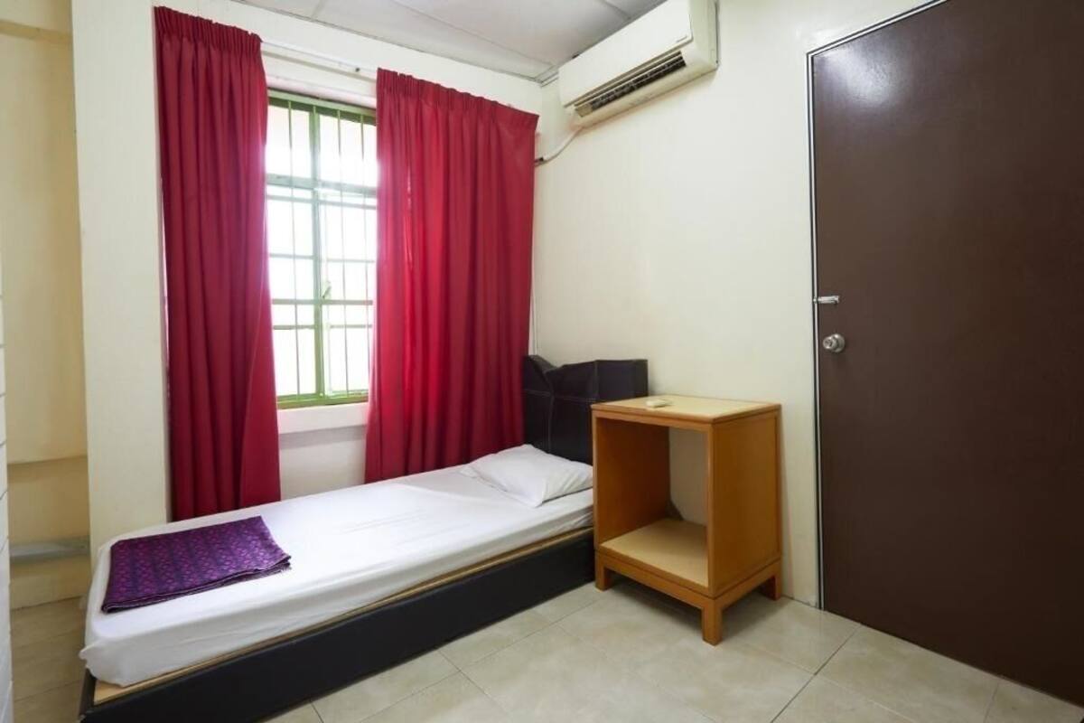 A small, single-bed room with a window, red curtains, a wooden nightstand, and a brown door.