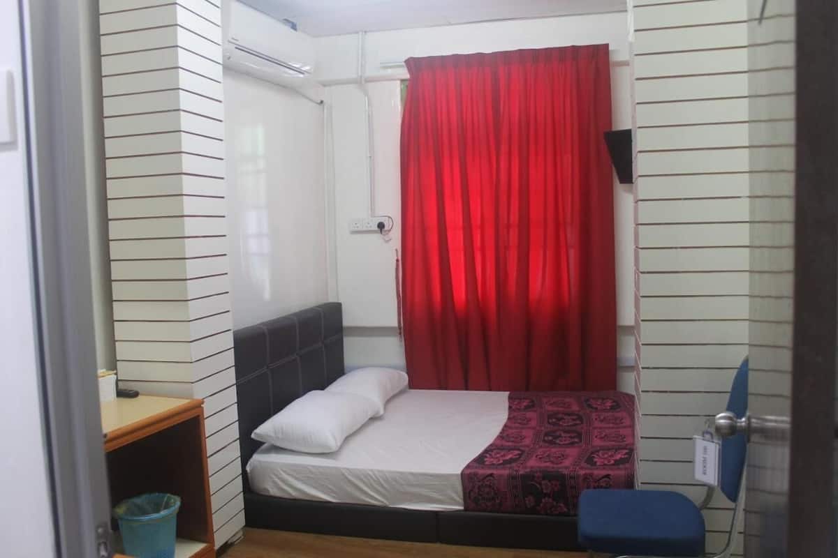 A small, compact room with a bed, a bedside table, a blue trash bin, and a red curtain.