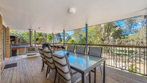 Une terrasse extérieure avec une table et des chaises en verre, un barbecue et une vue sur des arbres et un bâtiment.