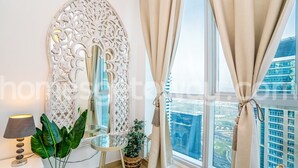 A modern room with a large ornate mirror, a potted plant, a small round table, and a view of a cityscape through large windows. - HomesGetaway-Torch Marina Dubai Cozy 2BR (Dubai)