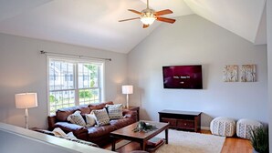 A living room with a brown leather sofa, a wooden coffee table, a TV mounted on the wall, and a ceiling fan.