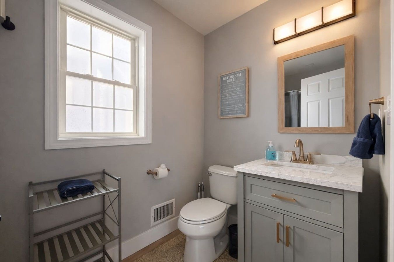 Un baño con inodoro, lavabo y espejo. Hay una ventana, un perchero para toallas y un cartel con normas.