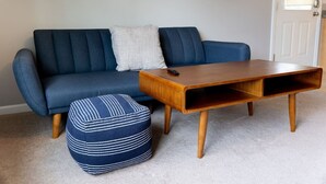 Un canapé bleu, une table basse en bois et un pouf à rayures se trouvent dans une pièce avec une porte blanche et un tableau encadré accroché au mur.