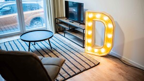 A living room with a television on a stand, a round coffee table, a large illuminated 'D' sign, and a view of the street through a window.