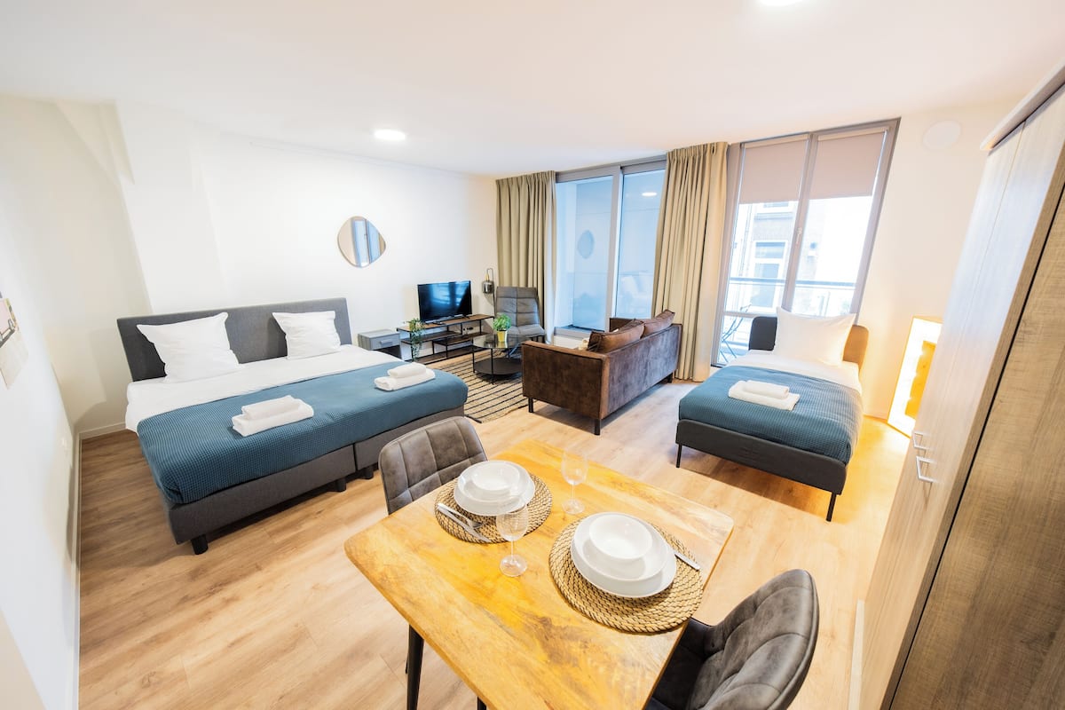A hotel room with a bed, a sofa, a dining table with plates and glasses, a TV, and a window with curtains.