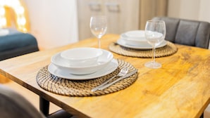 A wooden dining table with white plates and wine glasses, set with woven placemats and silverware.