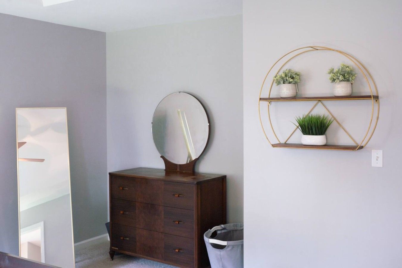 A room with a dresser, a mirror, a trash can, and a wall-mounted shelf with plants.