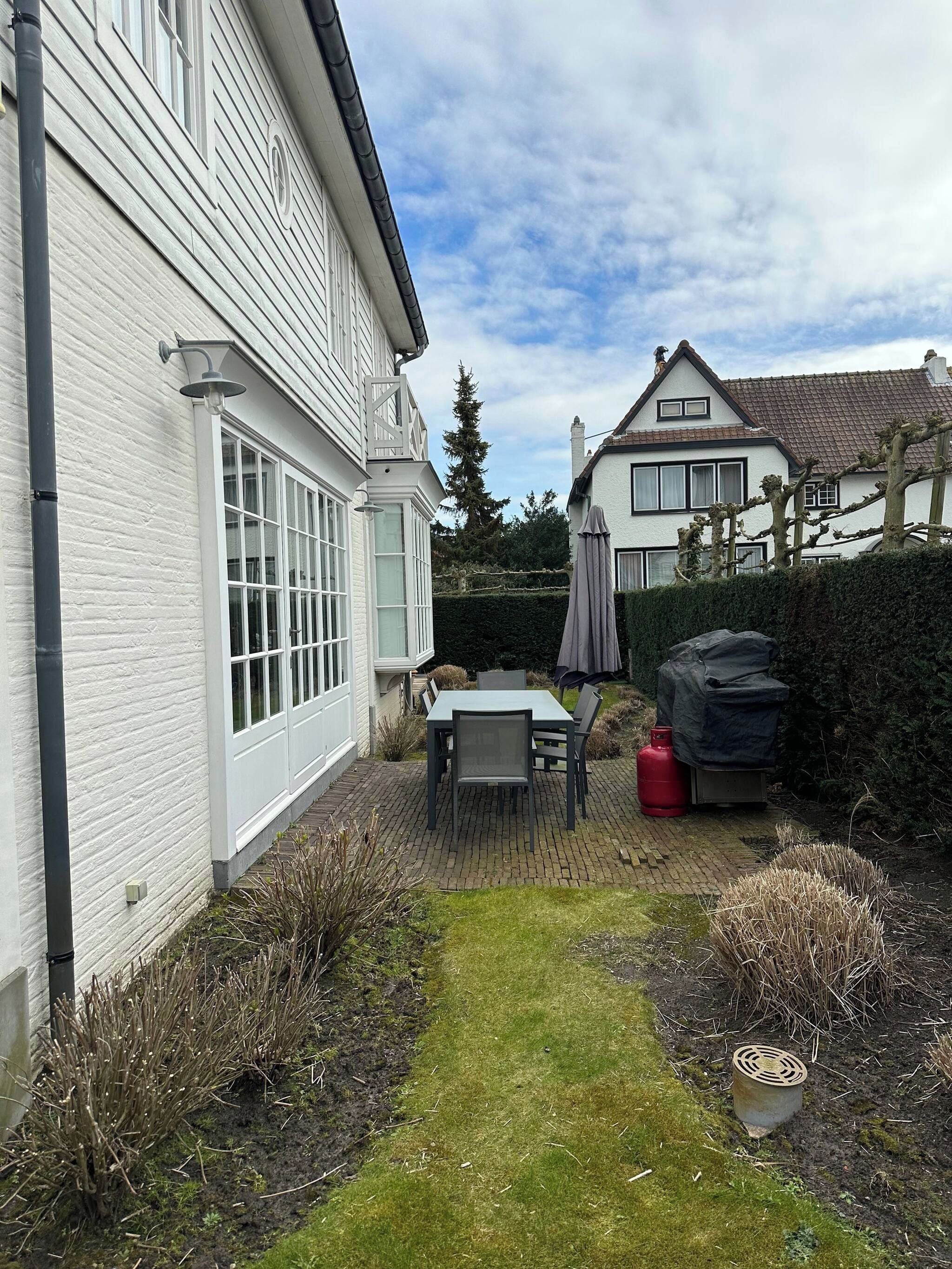 Een achtertuin met terras, een tafel en stoelen, een overdekte barbecue en een haag.