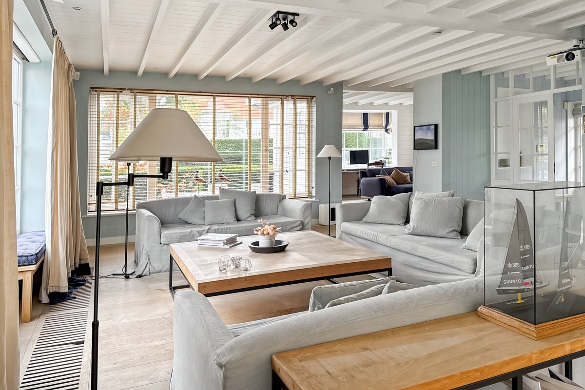Un vaste espace de vie comprenant un canapé, une table basse et une lampe. Une grande fenêtre, équipée de stores, offre une vue sur la verdure environnante.