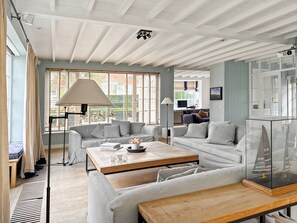 A spacious living area with a sofa, coffee table, and lamp. There is a large window with blinds and a view of greenery outside. - Luxurious 5-bedroom Holiday House in Knokke-heist Belgique (Knokke-Heist)