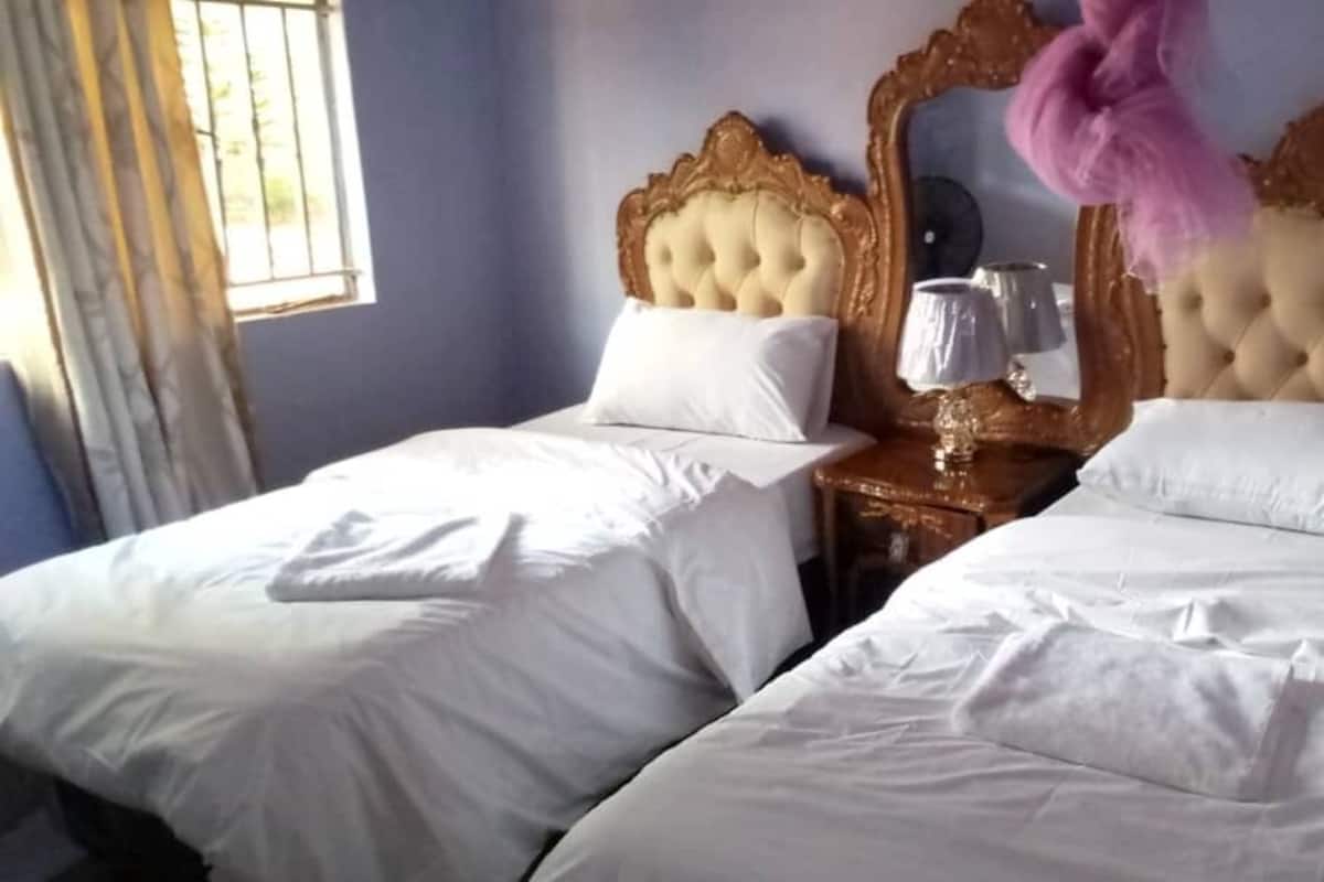 A room with two beds, each with a white bedspread and a single pillow. There is a wooden nightstand with a lamp and a mirror between the beds.