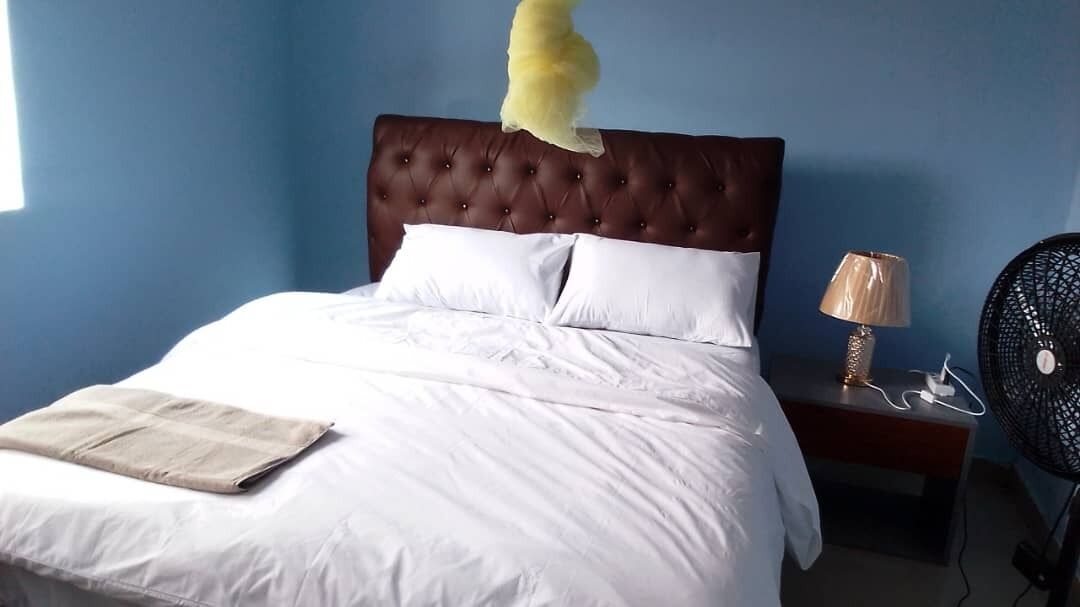 A bedroom with a tufted headboard, white bedding, a bedside table with a lamp, and a standing fan.
