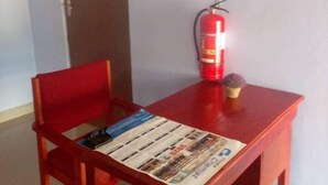 A small room with a red chair, a wooden table with a newspaper, a fire extinguisher, and a door marked "Room to Rooms". - Zambia (Chipata)