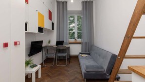 Un espacio habitable pequeño con un sofá gris, una mesita con una planta, un escritorio con una computadora portátil y un televisor montado en la pared.