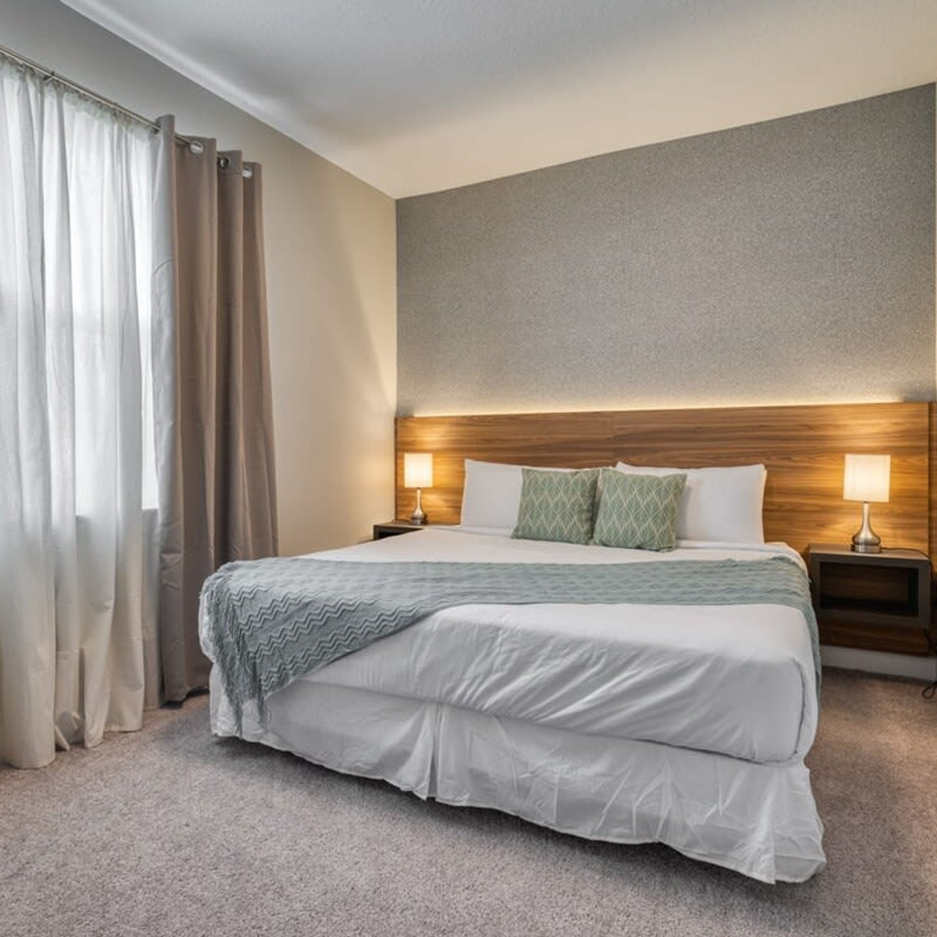 Quarto de hotel com uma cama grande, duas mesas de cabeceira com luminárias, uma cabeceira e vista para o exterior através de cortinas.