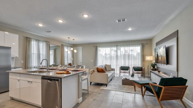 Una cocina moderna con una isla central, un comedor con sofá y sillas, y una sala de estar con televisor y asientos.