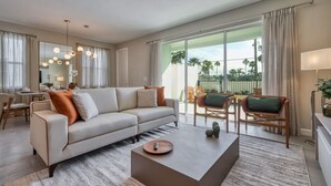Una sala moderna con un sofá, una mesa de centro y sillas. Hay un comedor con una mesa y sillas. Amplias ventanas ofrecen vistas a palmeras y una valla.