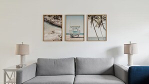 A grey sofa with two lamps on either side, a white wall, and three framed beach pictures. - Charming Townhouse Near Indialantic Beach (Indialantic)