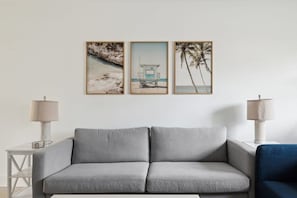 A living room with a grey sofa, two side tables with lamps, and three framed beach-themed pictures on the wall. - Charming Townhouse Near Indialantic Beach (Indialantic)