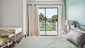A bedroom with a bed, a desk, a chair, a lamp, and a view of a balcony with a railing and greenery. - Charming Townhouse Near Indialantic Beach (Indialantic)