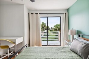 Ein Schlafzimmer mit Bett, Schreibtisch und Blick auf einen Balkon mit Geländer und Grünpflanzen.