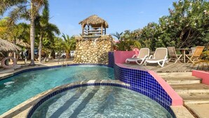A pool area with a stone wall, lounge chairs, and a thatched-roof structure. - Casa de las Brisas with Large Communal Pool (Kralendijk)