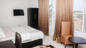 A hotel room with a bed, a chair, a small table, a TV, and a window with curtains. - Rivera Hotel and Suites (Ijede)