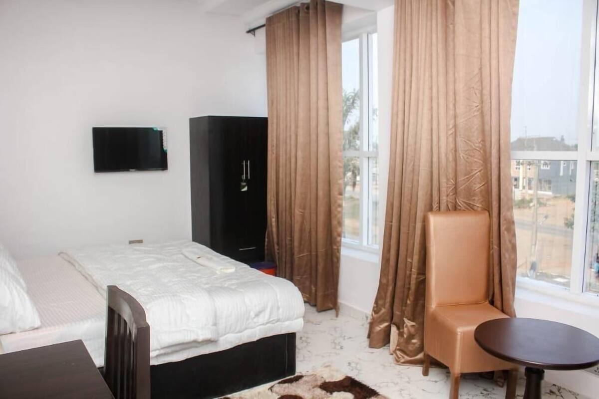 A hotel room with a bed, a chair, a small table, a TV, and a window with curtains.
