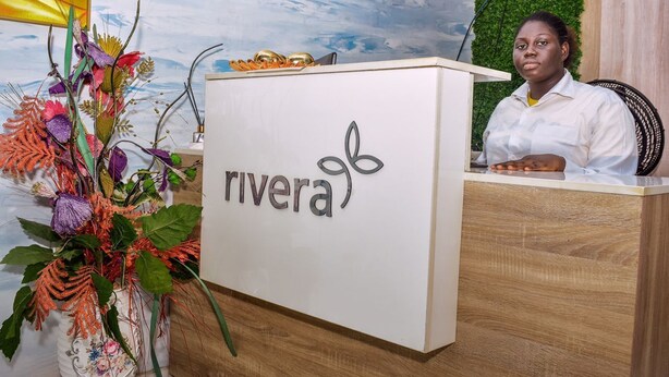 A reception desk with a logo, a decorative plant, and a green wall with a TV screen. - Rivera Hotel and Suites (Ijede)