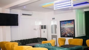 A room with a black leather sofa, yellow and teal chairs, a flat-screen TV, an air conditioning unit, and a painting on the wall. - Rivera Hotel and Suites (Ijede)