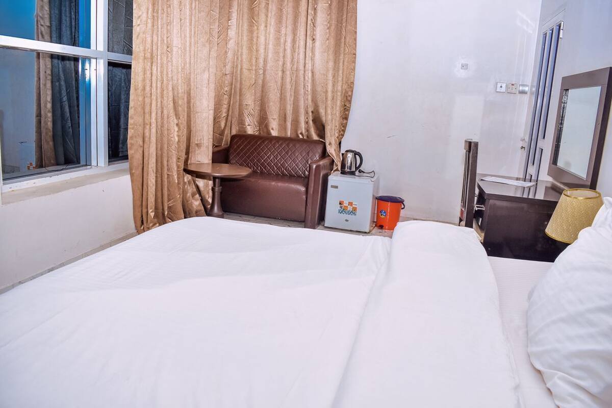 A hotel room with a bed, a small table, a chair, a minibar, a mirror, and a window with curtains.