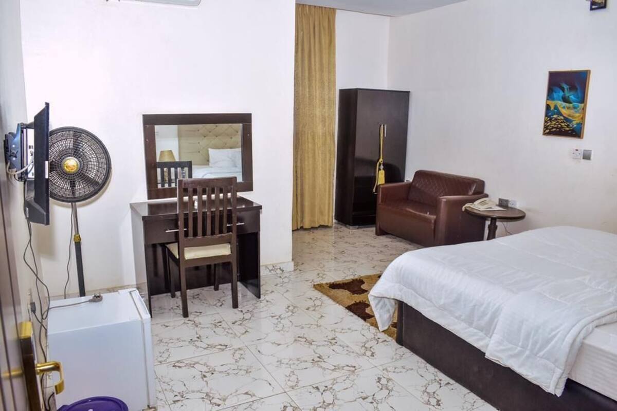 A hotel room with a bed, a desk with a chair, a television, a fan, a wardrobe, and a painting on the wall.
