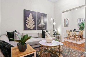 A modern living room with a sofa, coffee table, and decorative plants. There is a dining area with a table and chairs.