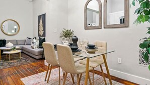 A modern living room with a glass dining table, beige chairs, a grey sofa, and a large mirror on the wall. - Spacious 3-Bed Brownstone, near Columbia Uni (New York)