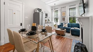 Una sala de estar moderna con una mesa de comedor de vidrio, un sofá azul y una cocina con armarios blancos y un refrigerador gris.