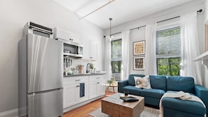 Una cocina moderna con armarios blancos, un refrigerador de acero inoxidable y un microondas. En la sala de estar, hay un sofá azul y una mesa de centro, con un ventanal que ofrece vistas a la vegetación exterior.