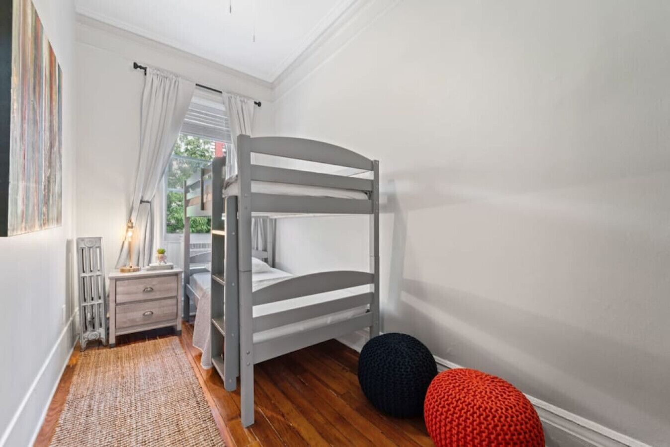 A room with a bunk bed, a small bedside table with a lamp, a red and black pouf, and a window with curtains.